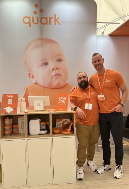 Die Mitbegründer von Quark, Senez und Gurinskas, stehen neben ihren Produkten an einem Messestand. Babyfoto auf der Rückseite mit ihrem Logo.